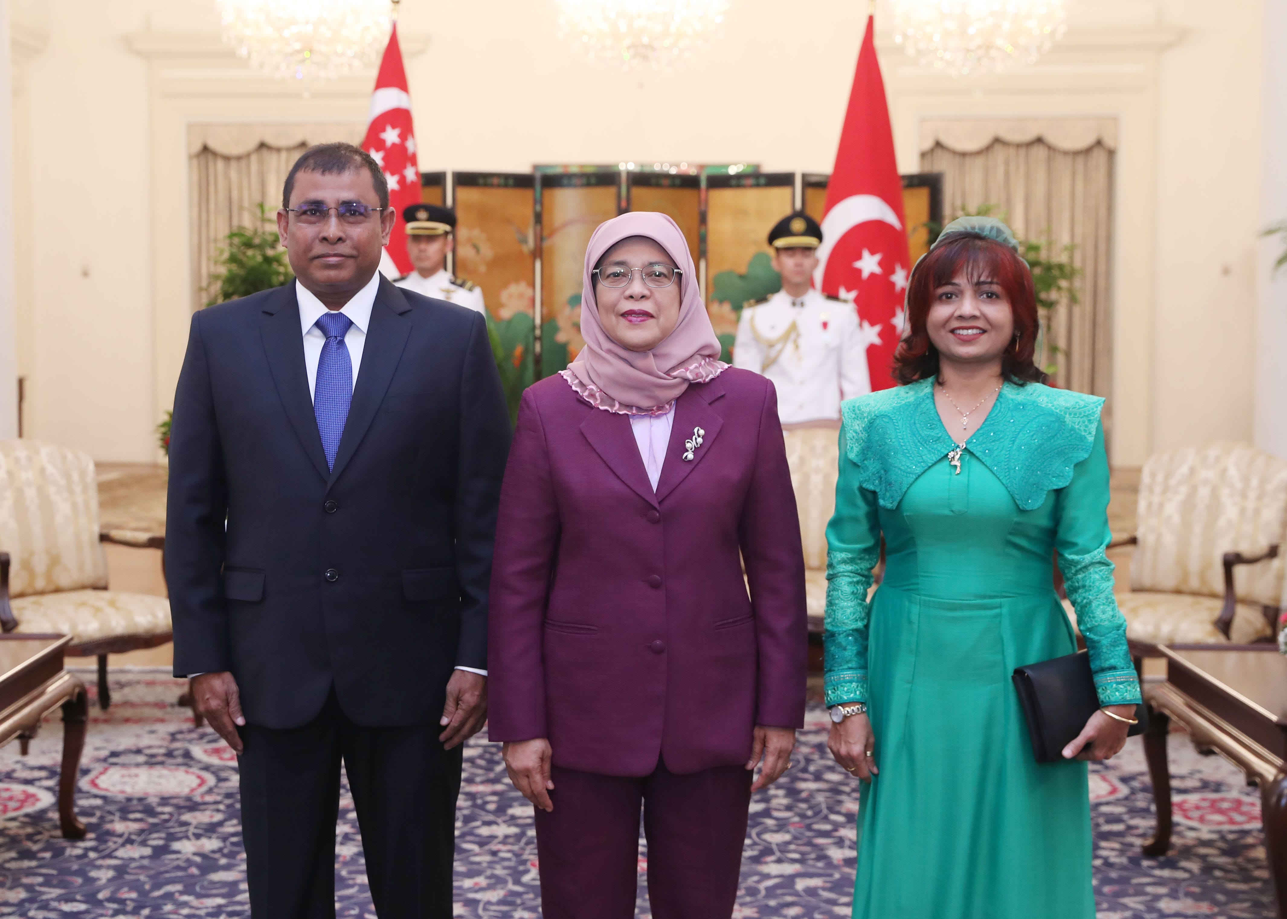 Three people stand before Singapore flags. Man in suit, woman in hijab, woman in green dress. Military personnel behind.